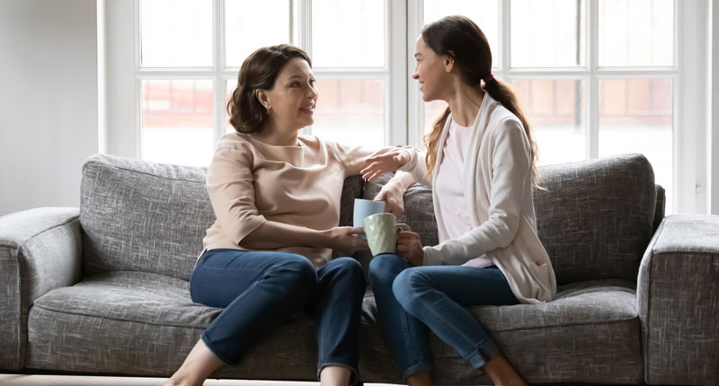 two women sit on a sofa in front of a large window, involved in conversation